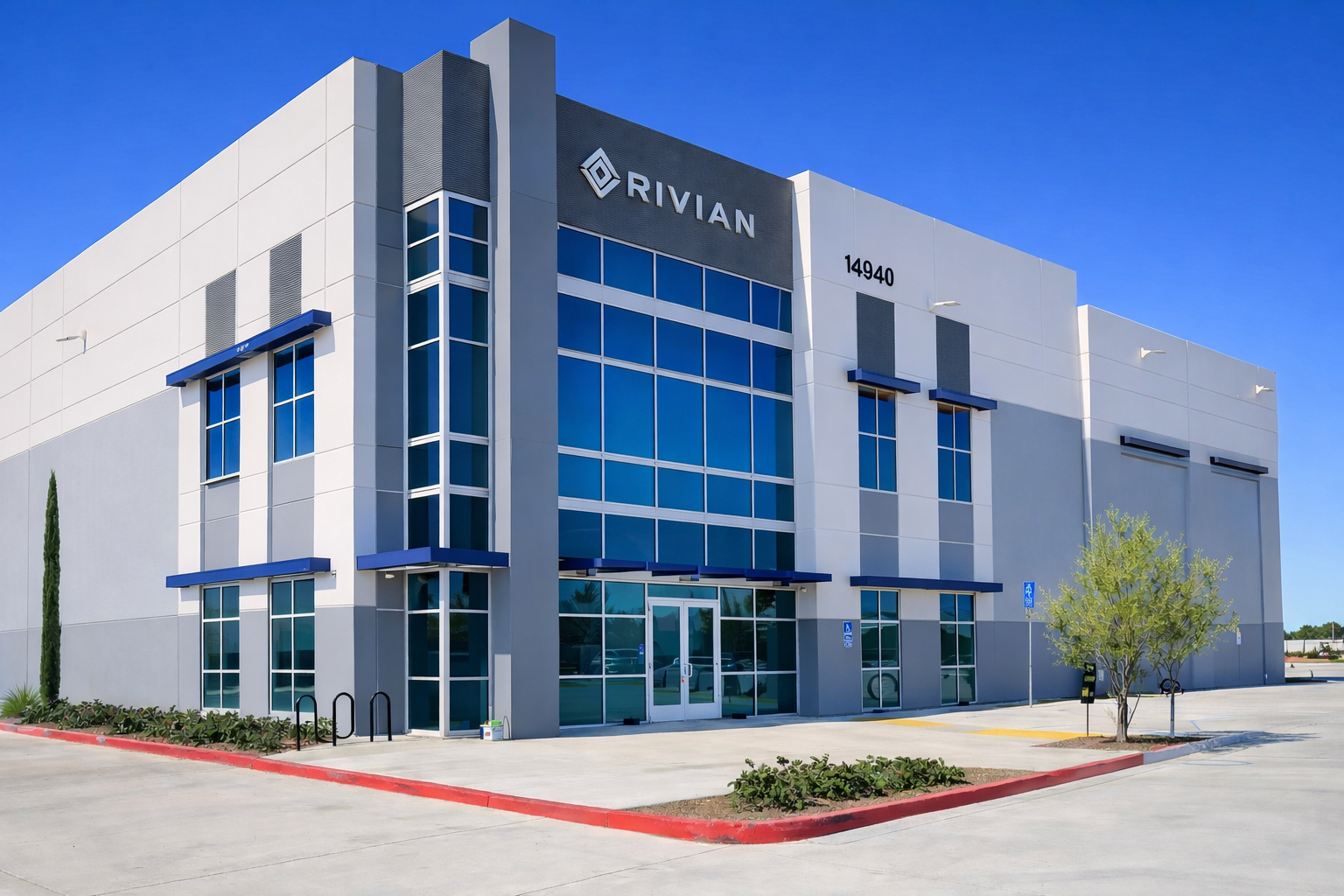 Modern commercial building with blue-tinted glass windows and the sign 'RIVIAN' on the front. Clear blue sky in the background, with a small tree and parking area in front. Structural engineering by BASE Design in San Francisco.