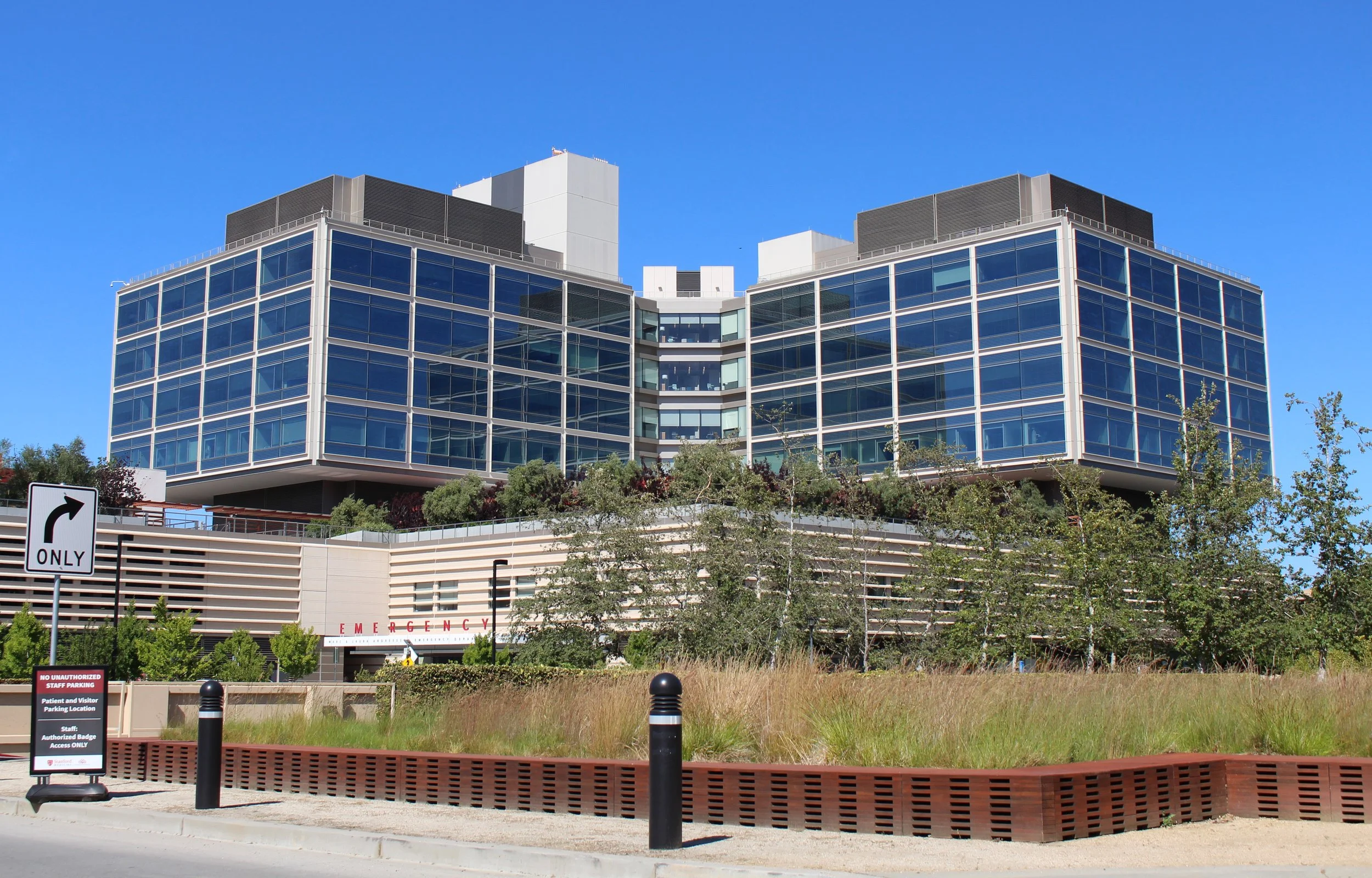 Stanford Hospital, a modern multi-story building with glass windows, surrounded by greenery and parking area, under a clear blue sky. Ongoing structural engineering support by BASE Design in San Francisco.