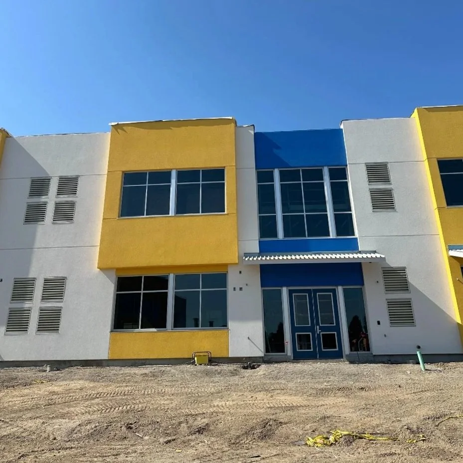 Under construction modern school property with white, yellow, and blue exterior, large windows, and the as yet unfinished dirt front yard under a clear blue sky. Structural engineering by BASE Design in San Francisco.