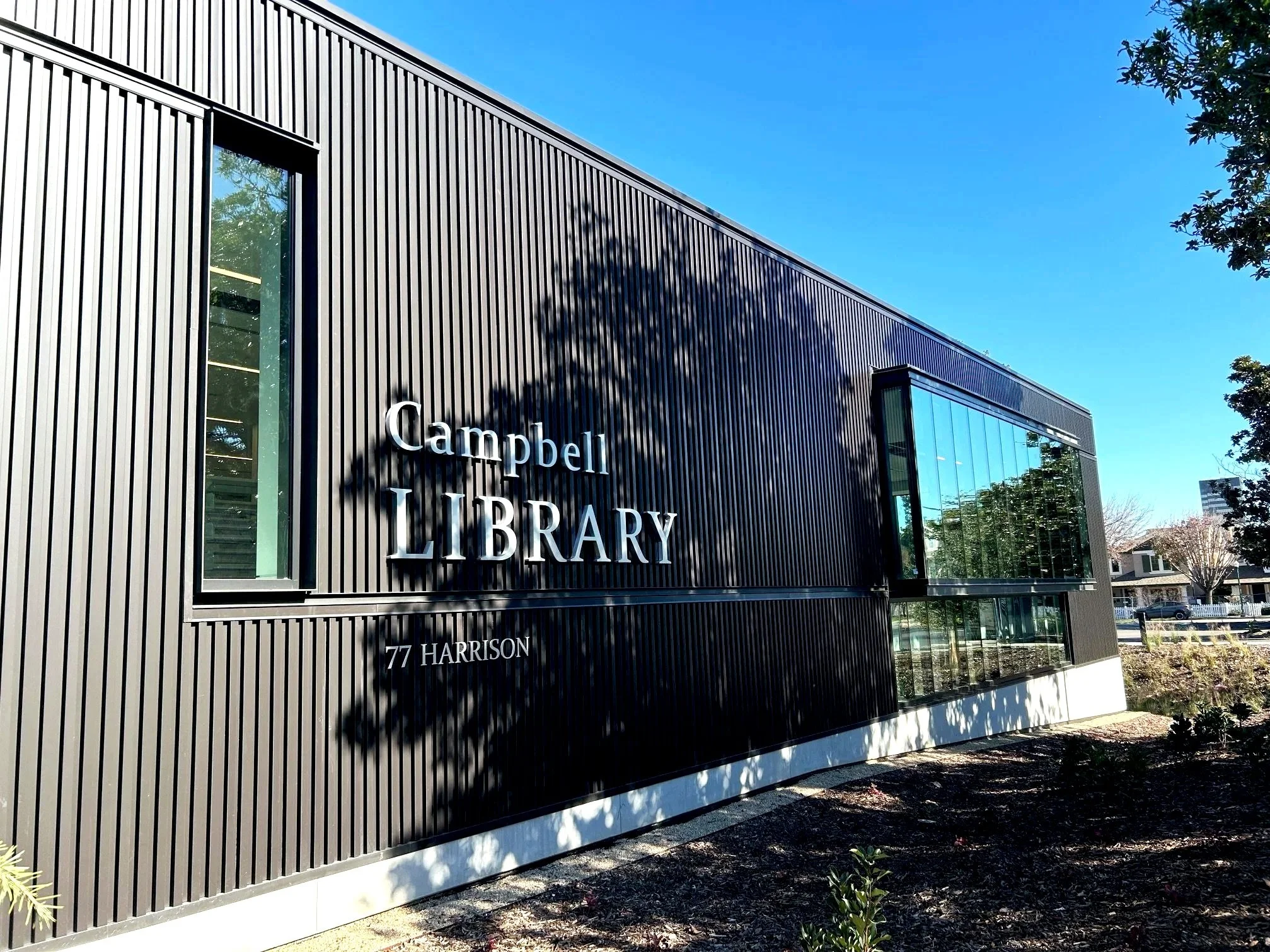 The exterior of the Campbell Library building, which has a modern design with black vertical panels and large glass windows. The library's name and address are displayed on the facade. Structural engineering by BASE Design in San Francisco.