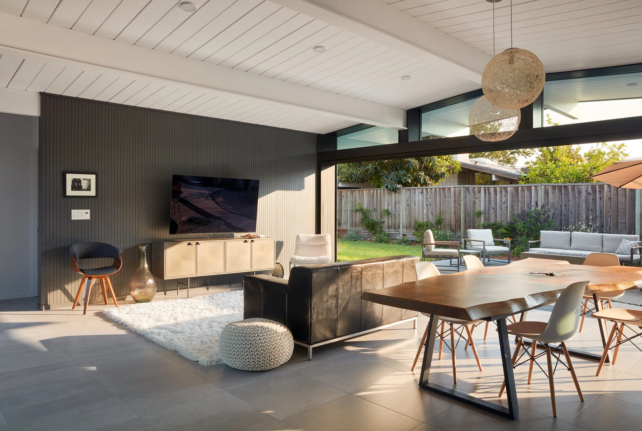 Living room of an Eichler-designed home in Palo Alto, CA, with large glass doors opening to backyard, modern furniture, flat-screen TV, wood dining table, chairs, and outdoor seating area. Structural engineering by BASE Design in San Francisco.