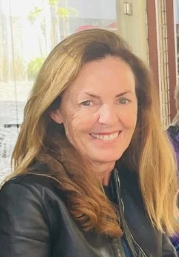 A woman with long, wavy blonde hair smiling indoors with a window and red brick wall behind her.