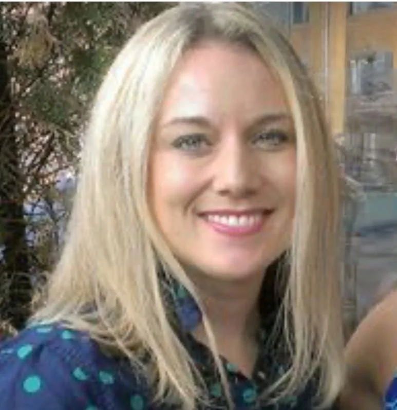 A smiling woman with long blonde hair, wearing a dark blue top with teal dots, outdoors with trees and a building in the background.