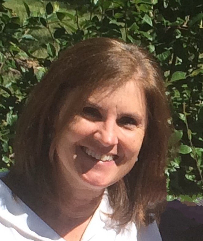 A woman with shoulder-length brown hair smiling outside in front of green bushes.