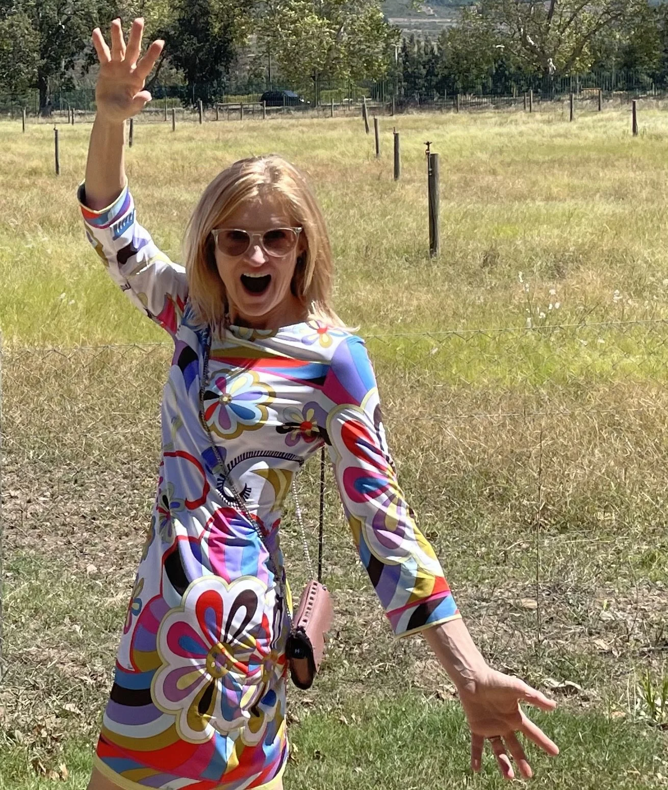 A woman in a colorful floral dress and sunglasses is outdoors in a field, smiling and waving with one hand while holding a small purse with the other.