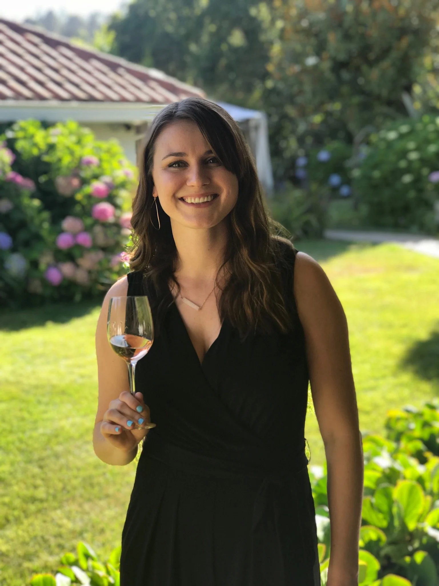 Smiling woman in a black dress holding a glass of rosé wine outdoors, with green garden and blooming flowers in background.