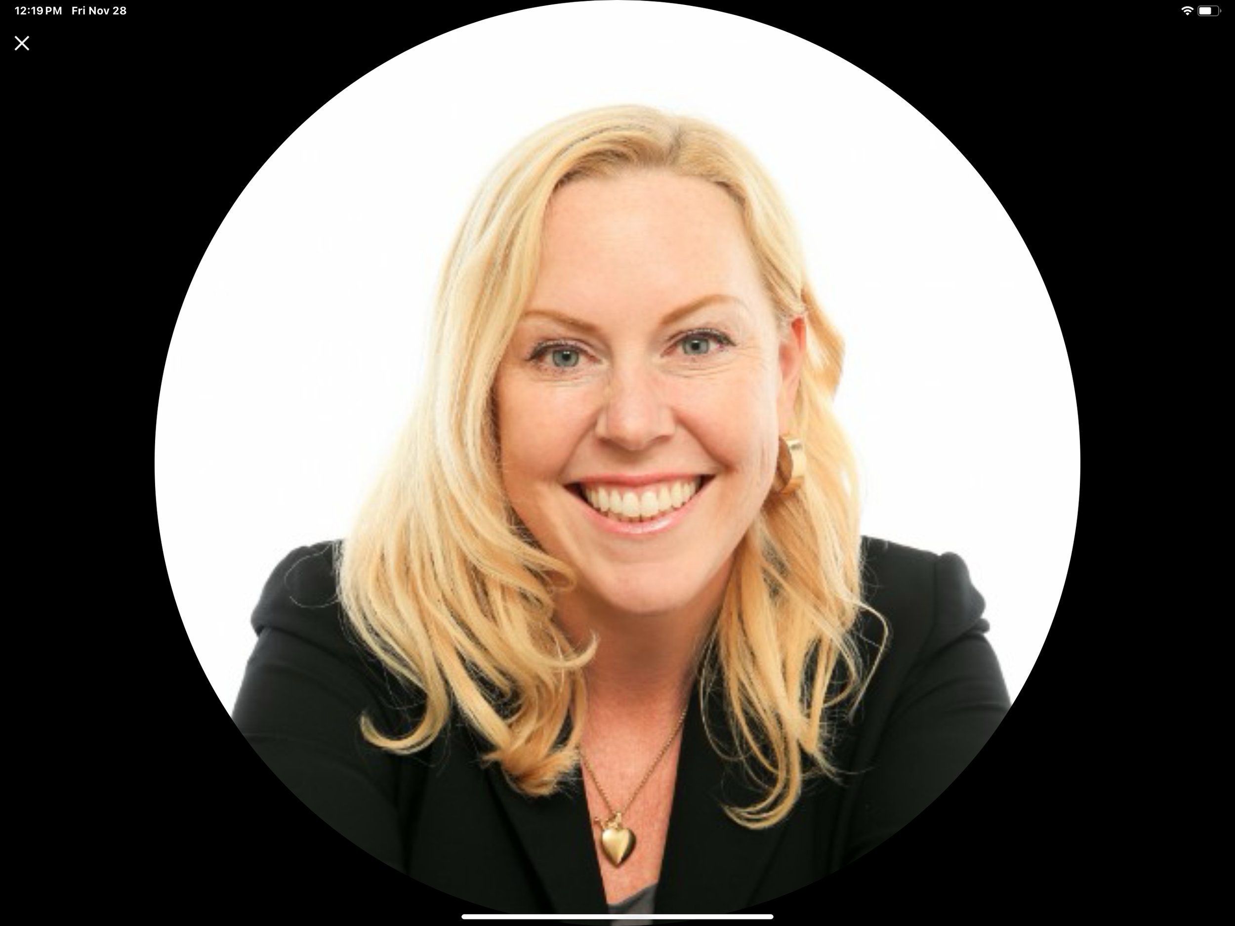 Portrait of a smiling blonde woman wearing a black blazer, gold necklace with a heart pendant, and large earrings, against a white background.