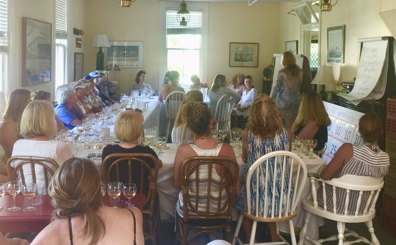 A group of women attending a workshop or meeting in a dining room with a long table, wine glasses, and a person standing at the front near a whiteboard.