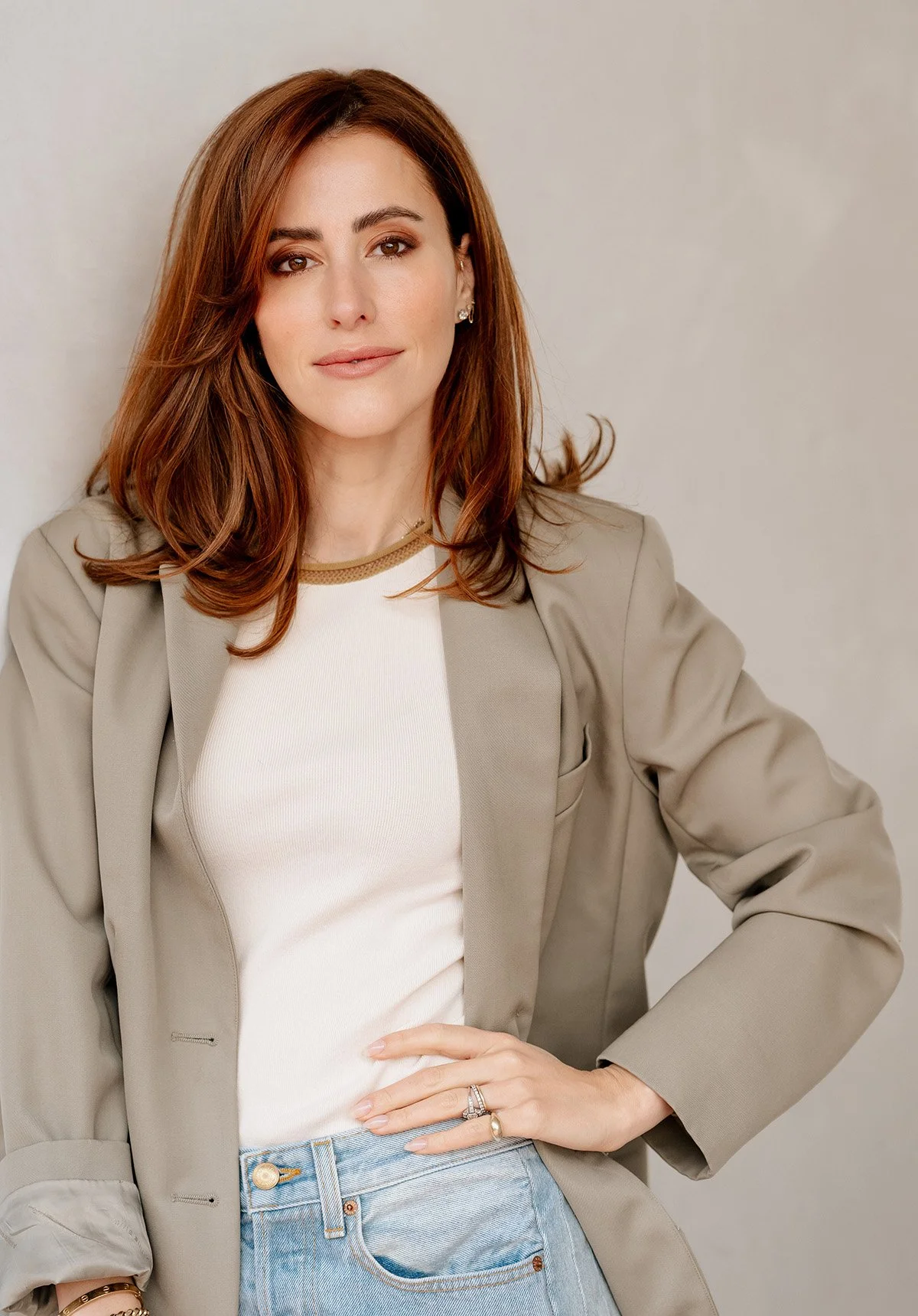 Dr. sarah Oreck with shoulder-length red hair, wearing a beige blazer over a white shirt and blue jeans, posing against a plain light-colored background.