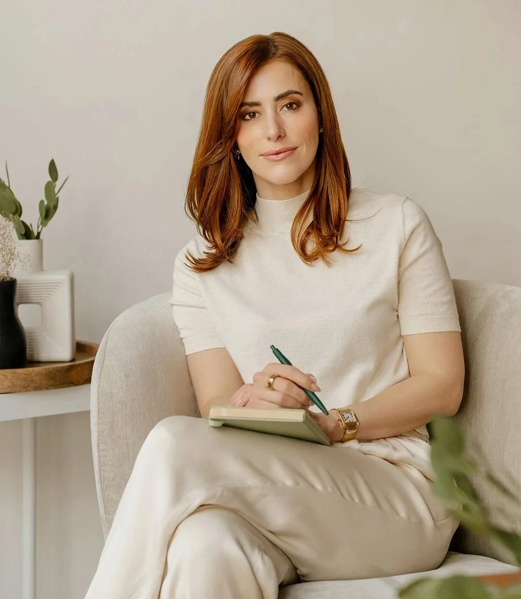 Dr. Sarah with medium-length auburn hair, wearing a cream-colored t-shirt and beige pants, sits on a light-colored sofa, holding a notebook and pen. She is in a cozy, well-decorated living space with a potted plant and a small table in the background