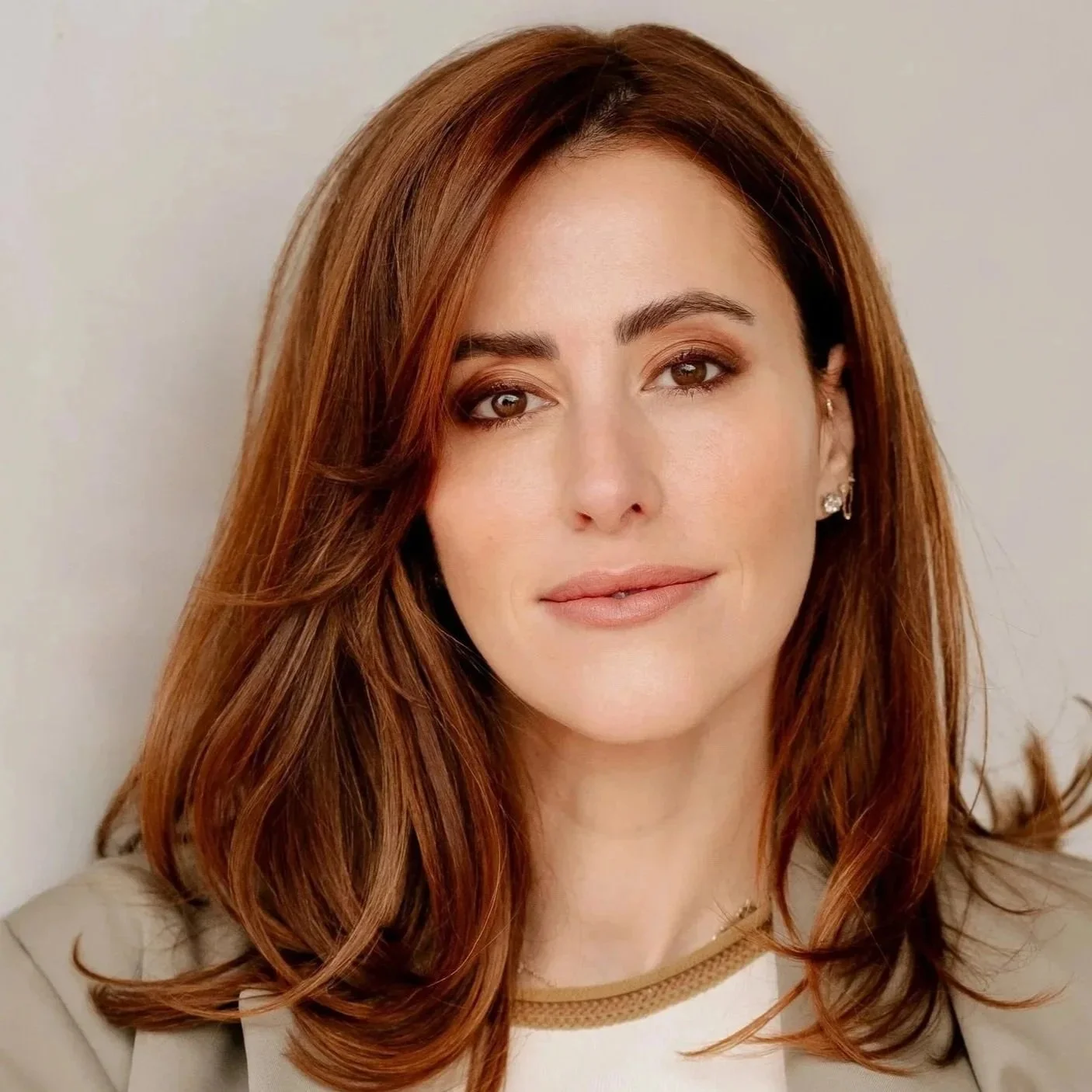 Portrait of Dr. Sarah Oreck with long, wavy auburn hair, light complexion, wearing makeup and earrings, against a plain background.