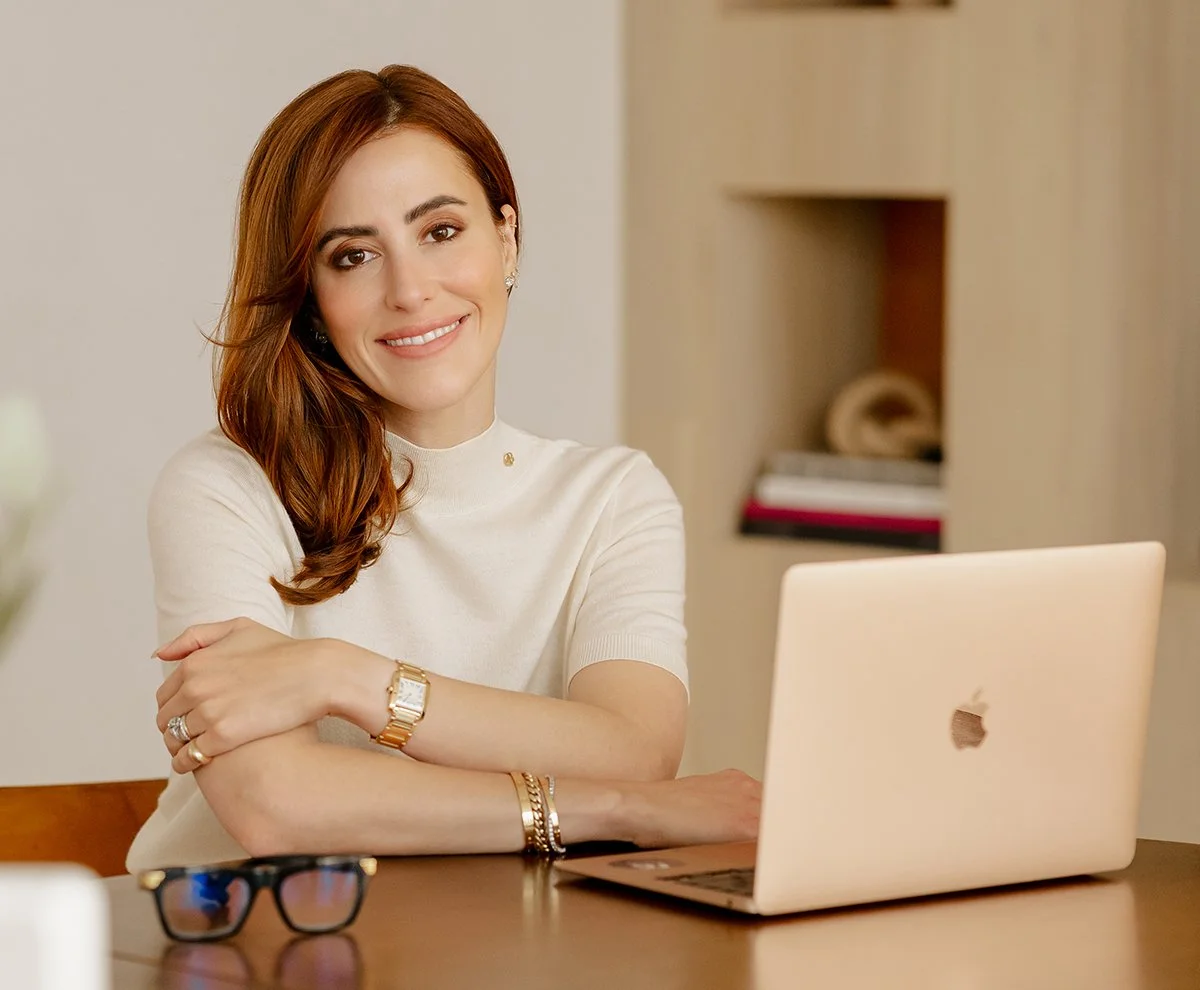 Dr. Sarah Oreck with wavy, reddish-brown hair and light skin sitting at a wooden desk with a silver Macbook, wearing a white short-sleeved shirt, and jewelry, smiling at the camera, with glasses on the desk.