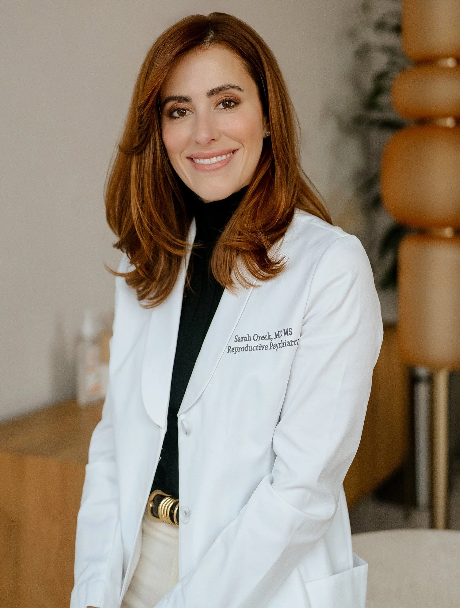 Dr. Sarah Oreck shoulder-length auburn hair, smiling, wearing a white lab coat over a black turtleneck, standing indoors.