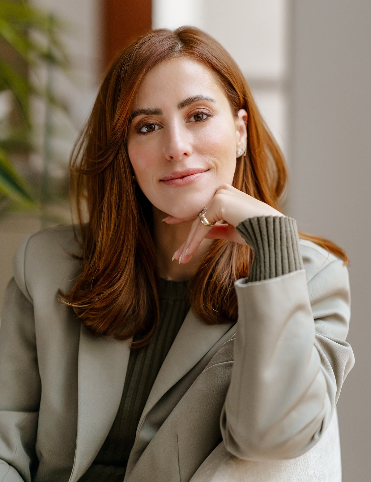 Dr. Sarah Oreck with red hair and light skin, wearing a beige blazer and a green top, resting her chin on her hand and smiling gently at the camera.
