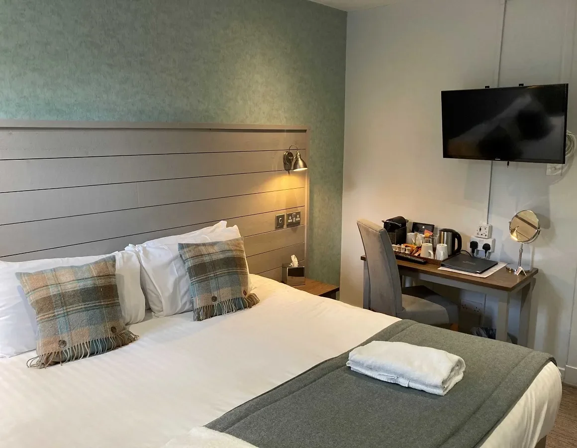 Hotel room with a bed, plaid pillows, wall-mounted lamp, TV, and a desk with tea/coffee supplies and a laptop.