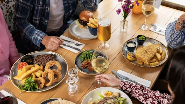 A group of people enjoying a meal together at a restaurant table with plates of fried fish, burgers, steak, onion rings, French fries, salads, and drinks, including wine and beer.