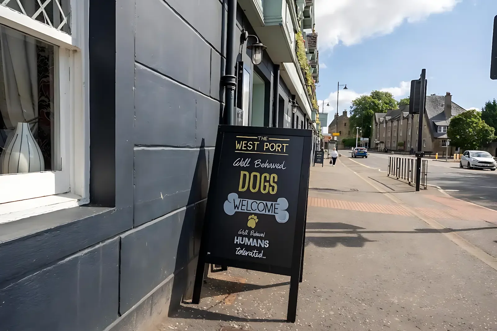 A sidewalk scene outside a restaurant with a blackboard sign welcoming dogs and humans, in a town with parked cars and historic buildings, bathed in sunlight.