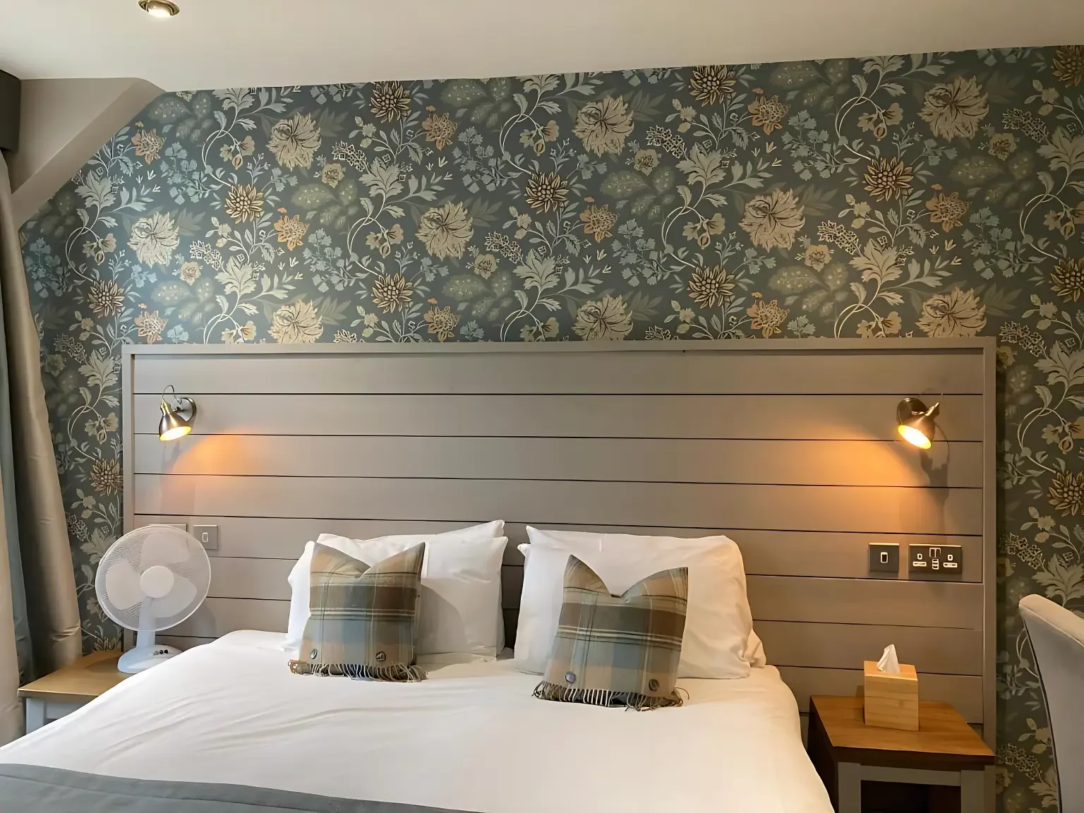 A hotel room with a bed featuring two plaid pillows, a wooden headboard with built-in reading lights, a small bedside table with tissue box, a white fan, and floral wallpaper in the background.