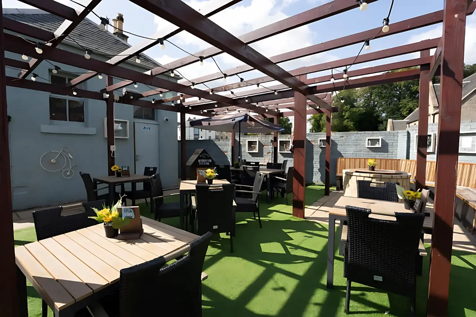 Outdoor patio with tables, chairs, string lights, and a brick fireplace under a pergola with a cloudy sky