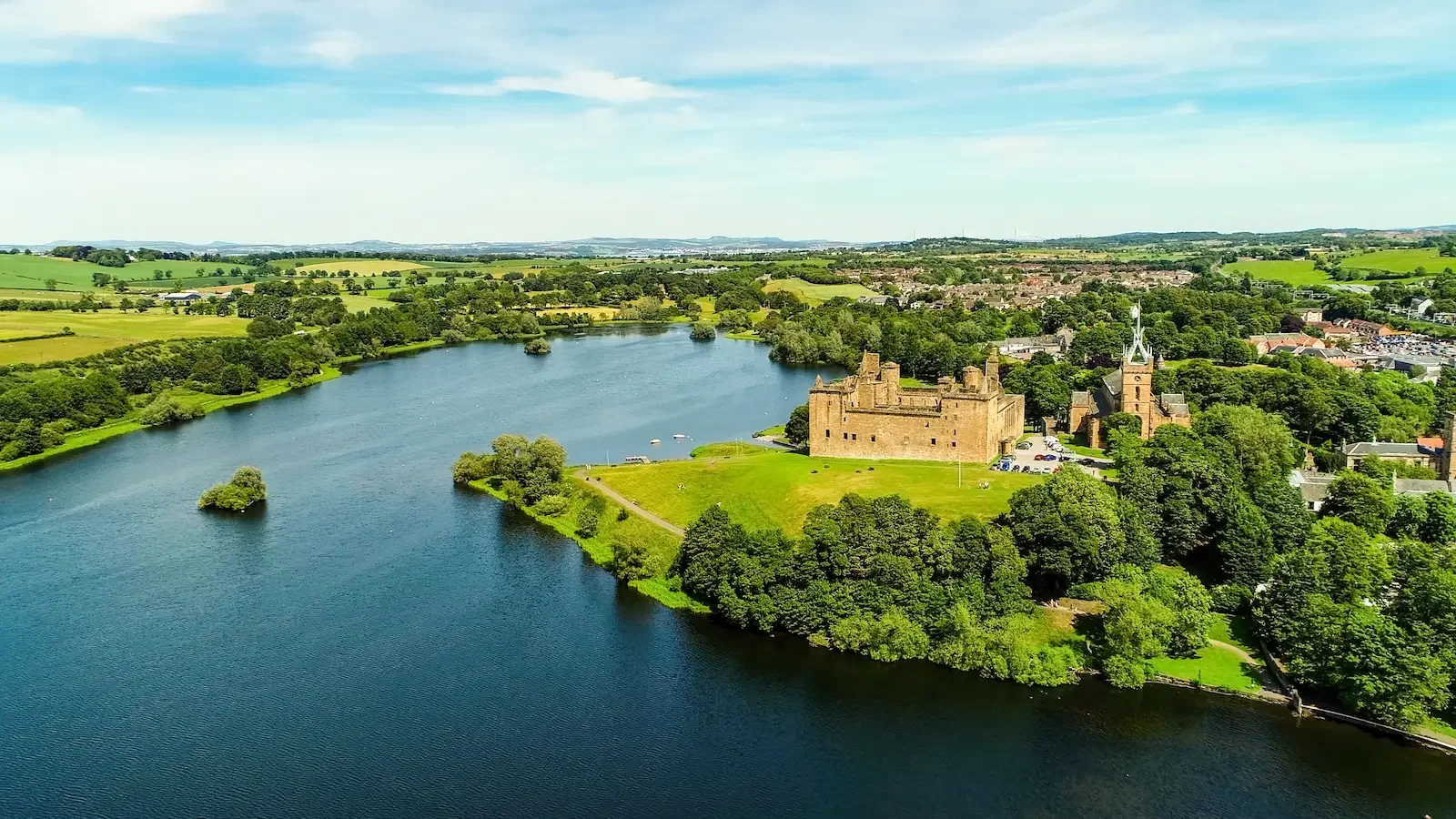 Aerial view of an old castle surrounded by a large body of water and lush green land with scattered trees and buildings, under a partly cloudy sky.