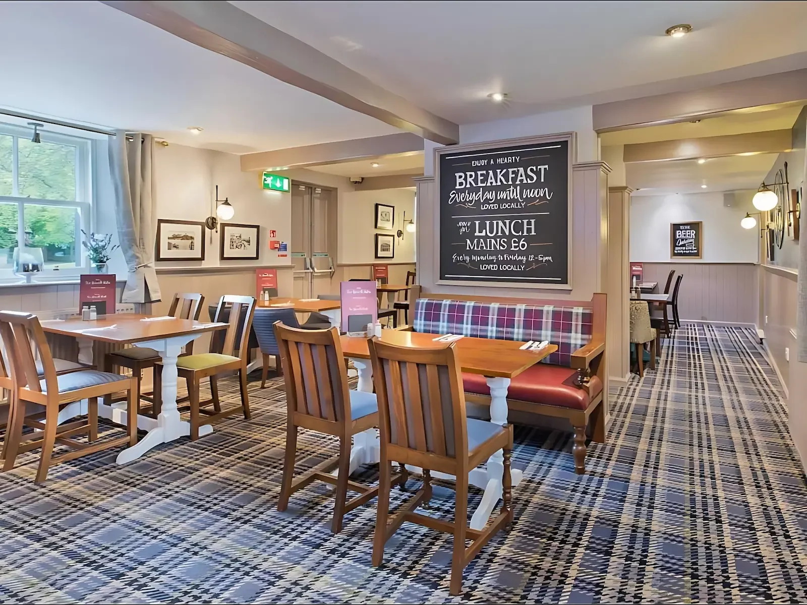 Interior of a cozy restaurant or cafe with wooden tables and chairs, checkered carpet, and a large blackboard menu displaying breakfast and lunch specials. The space is brightly lit with wall-mounted lamps and natural light from a window with curtains.