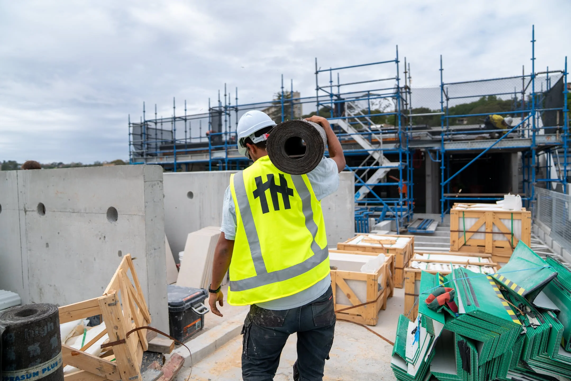 Construction worker wearing a yellow safety vest and white helmet, carrying a roll of material over his shoulder at a building site with scaffolding and construction supplies.