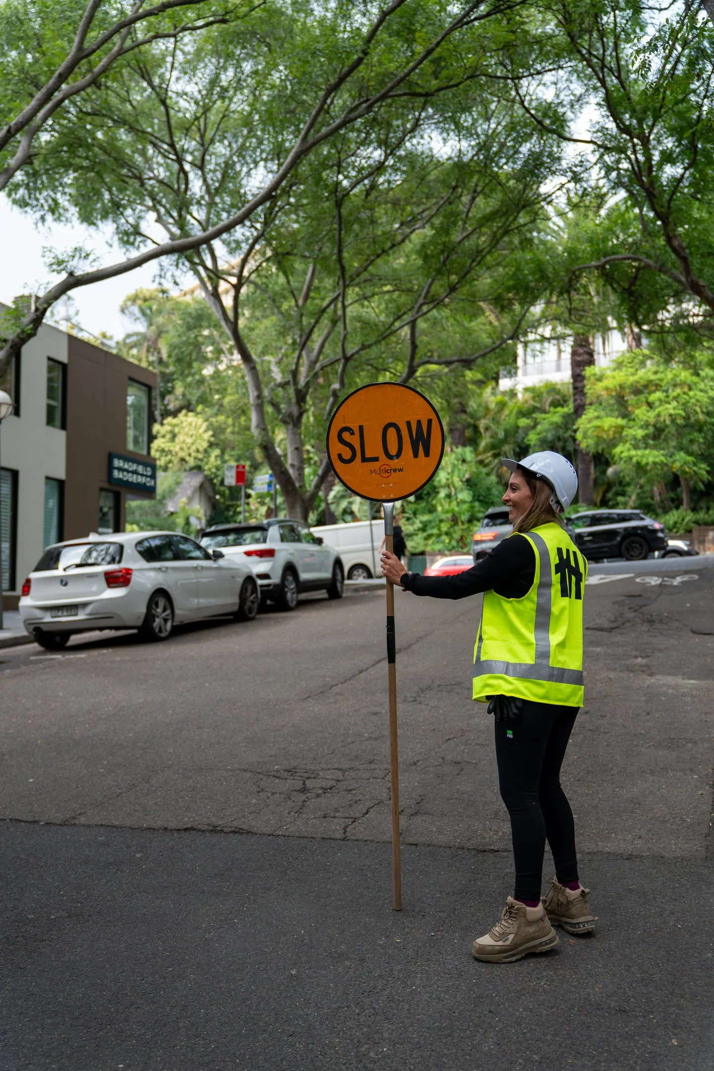 A woman wearing a safety helmet and yellow reflective vest holding a bright orange 'SLOW' sign while standing on a street lined with parked cars and green trees.