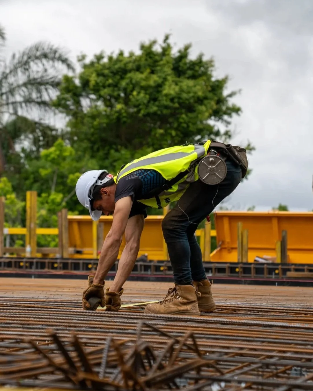 Construction worker wearing a white safety helmet, yellow vest, and brown boots bending down on a steel rebar grid at a construction site outdoors.