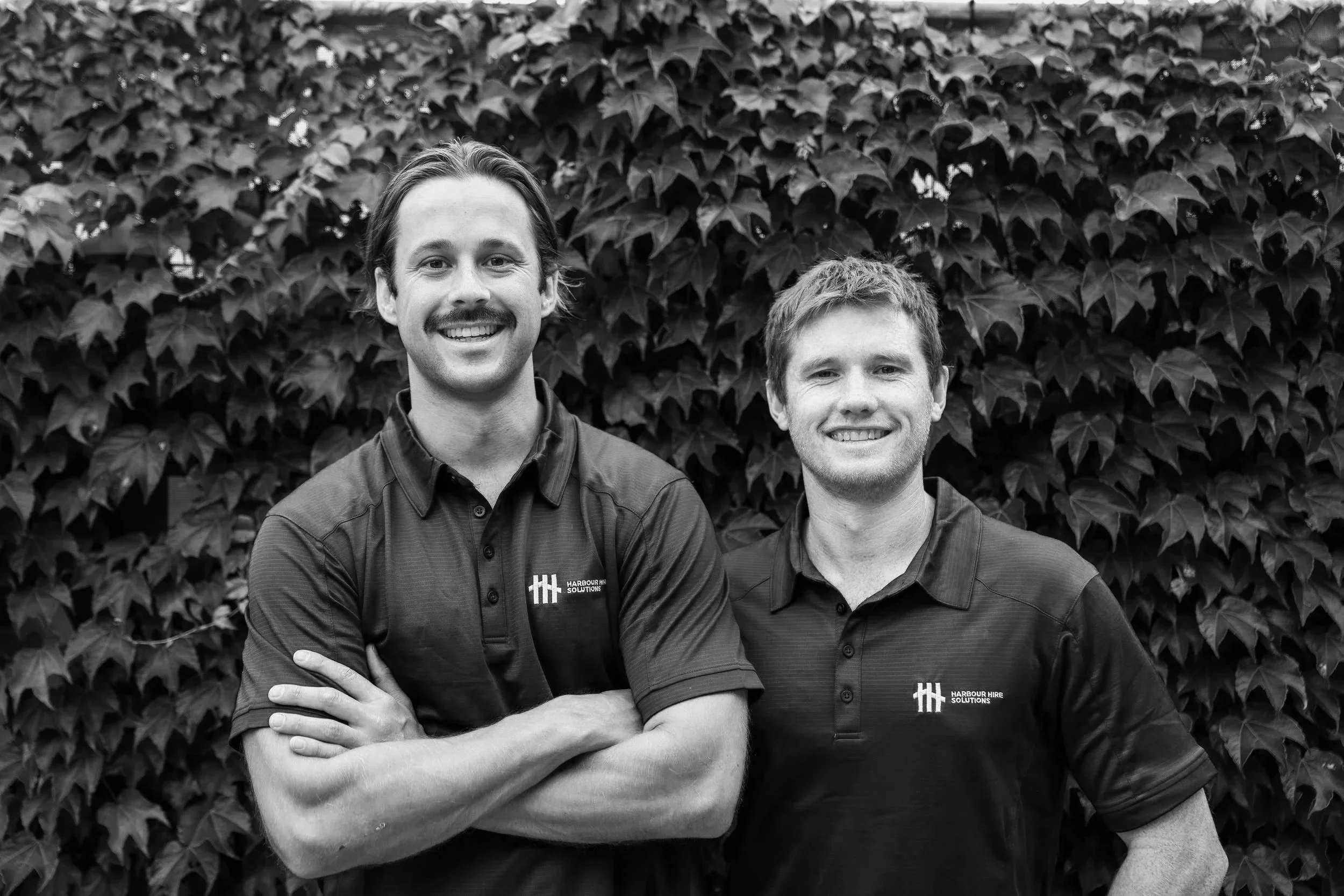 Two smiling men with arms crossed standing in front of a leafy hedge, wearing collared shirts with 'Harbour Hire Solutions' logos.