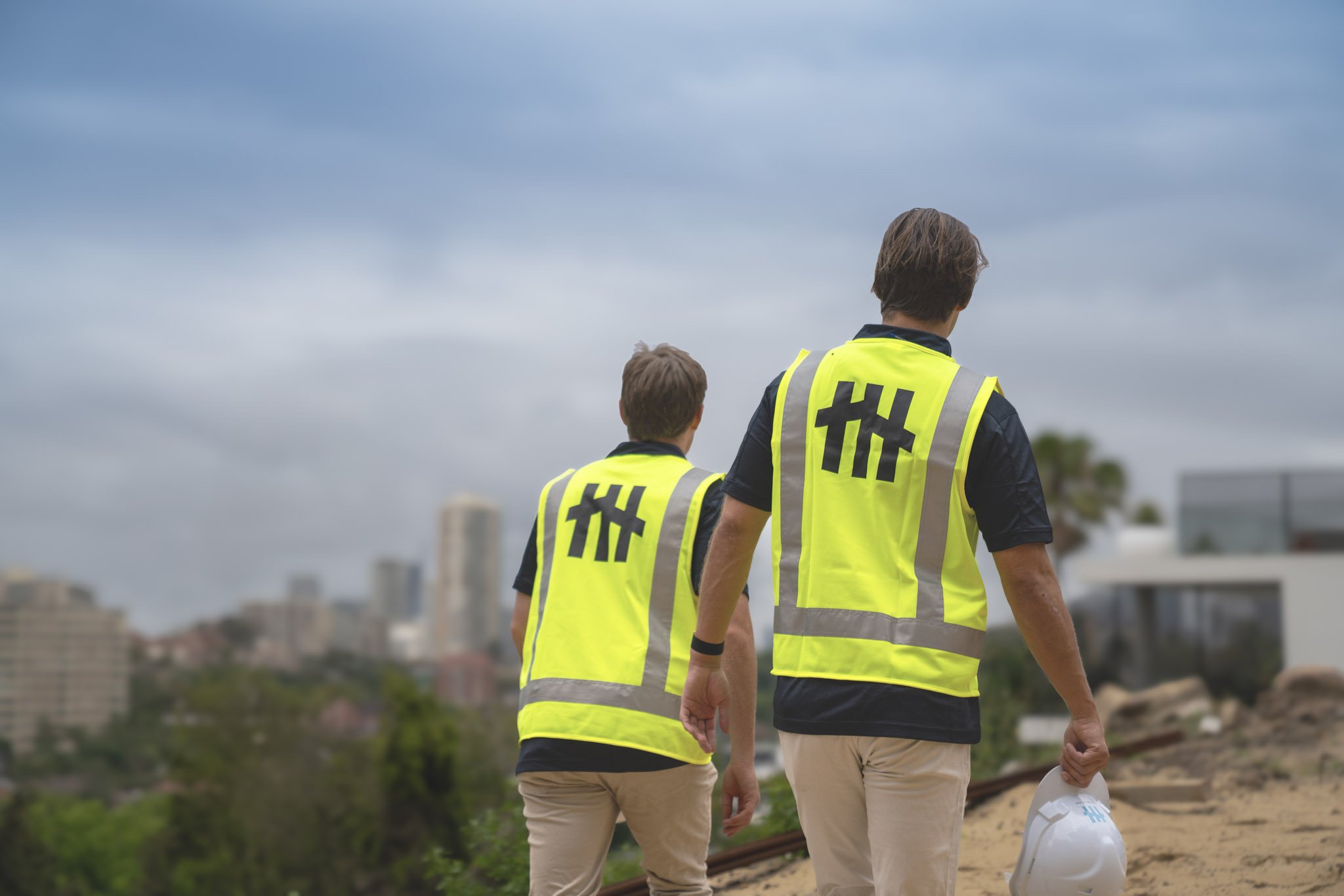 Two construction workers wearing yellow safety vests walking on a dirt path near railway tracks, with a city skyline and cloudy sky in the background.