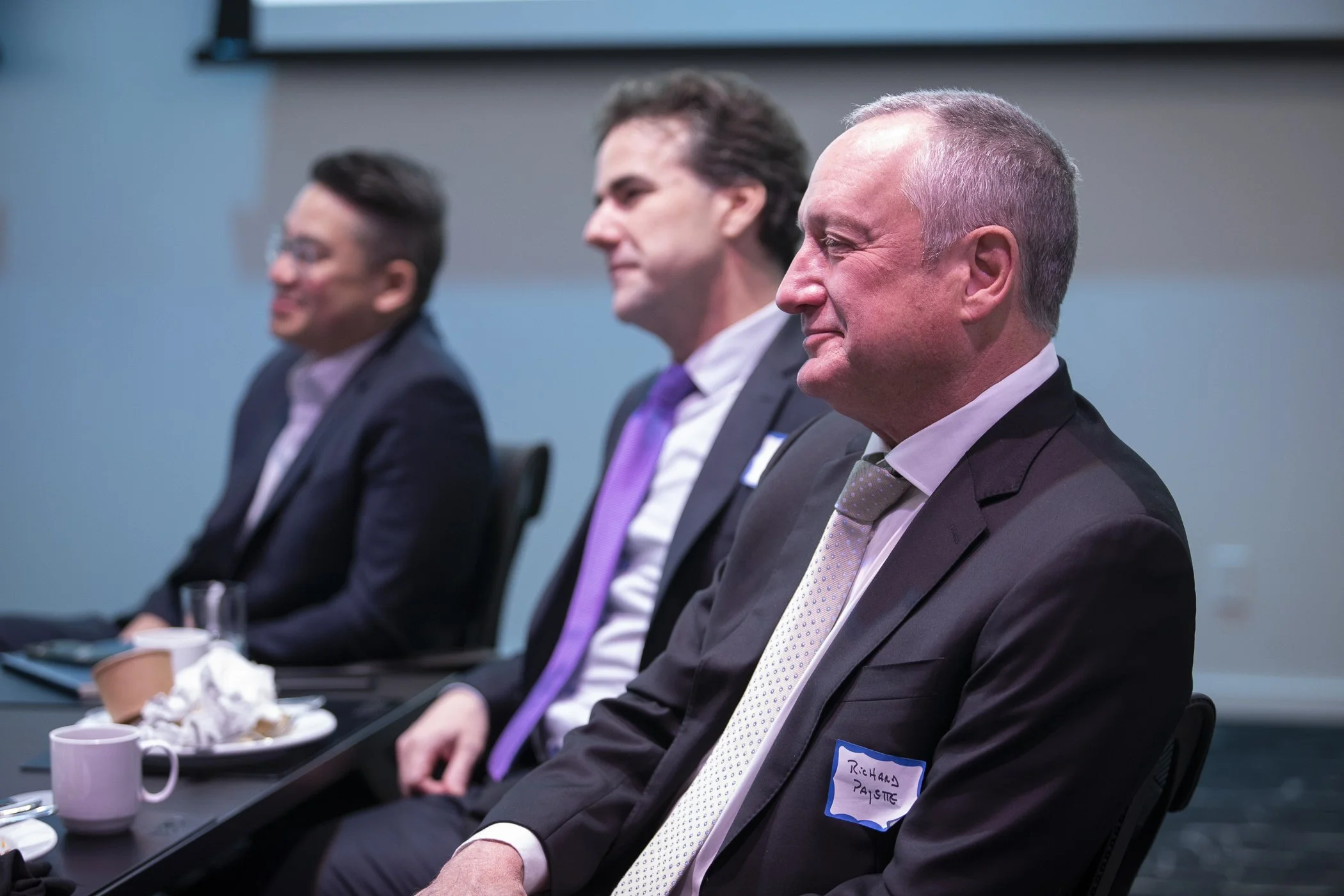 Three men in business suits sitting in a row at a conference or meeting, with cups and plates on the table in front of them.
