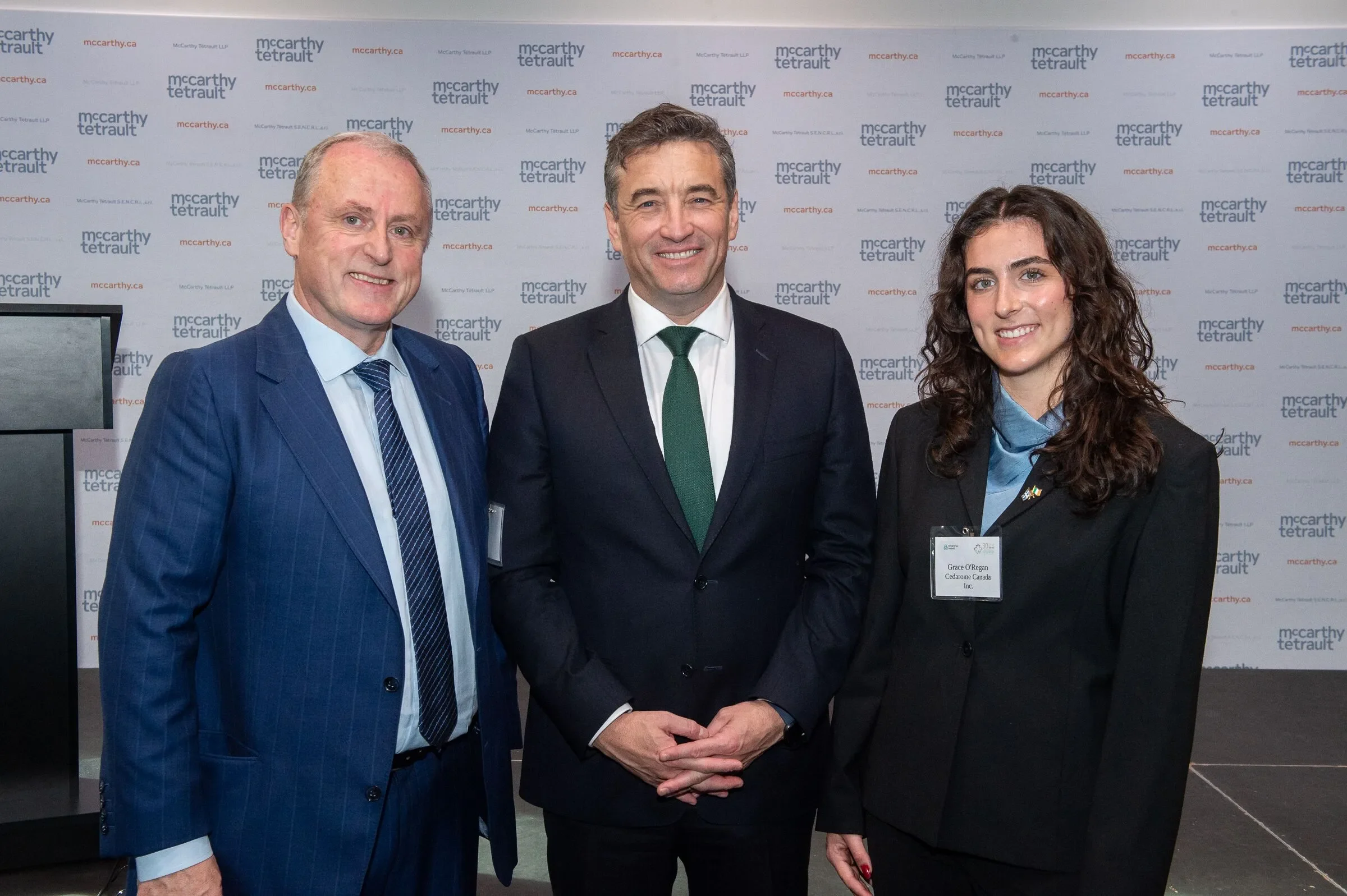 Three professionally dressed individuals standing together at an event, posing for a photo with a branded backdrop that says 'mccarthy tetrault' repeated in the background.
