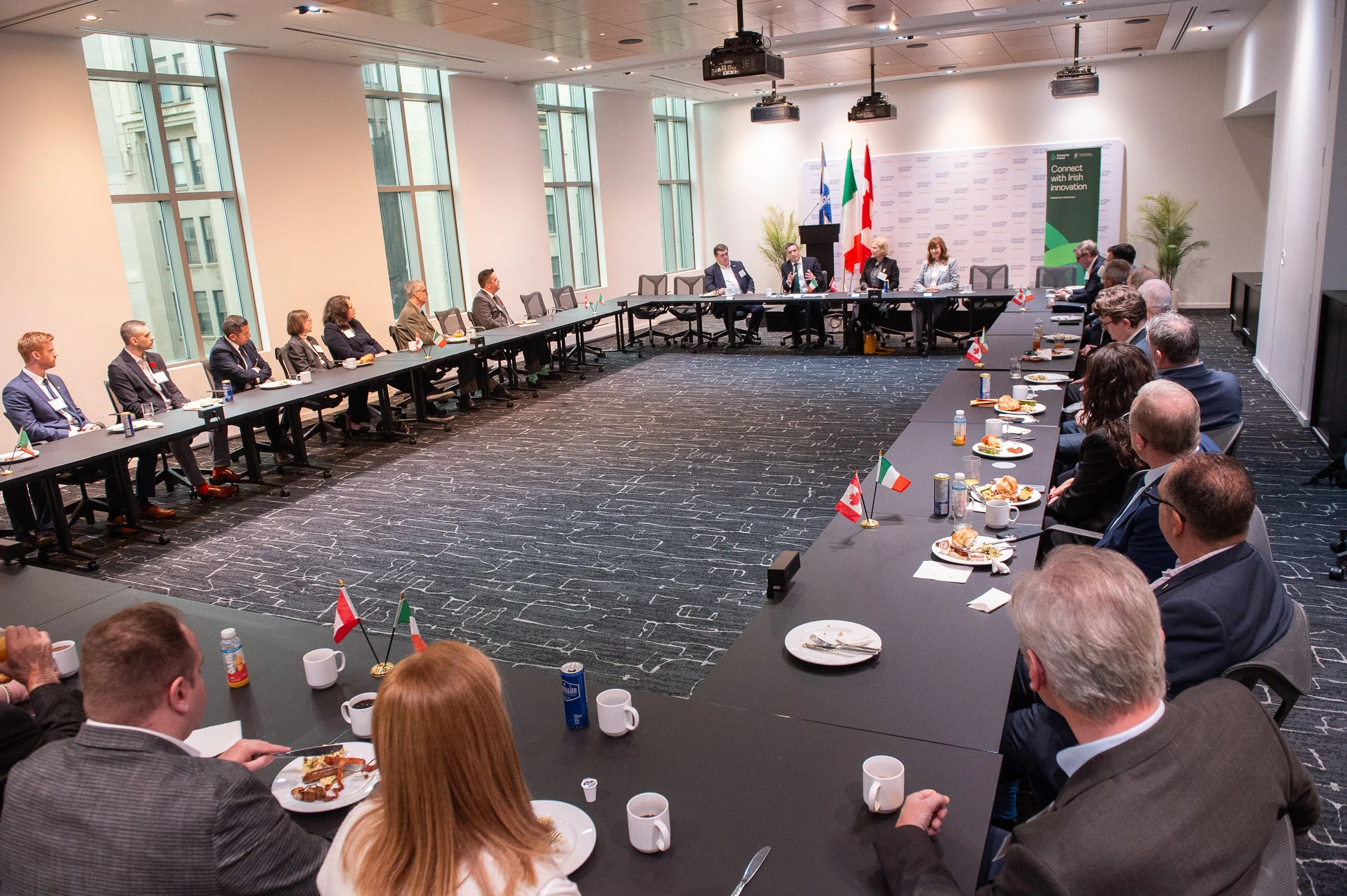 A large conference room with a U-shaped table setup. There are about 20 people seated, dressed in business attire, some with small flags of Italy, Canada, Ireland, and the United Kingdom on the table. Four individuals are seated at the head of the room, with flags, microphones, and a backdrop bearing logos behind them. The room has high windows, a dark patterned carpet, and potted plants.