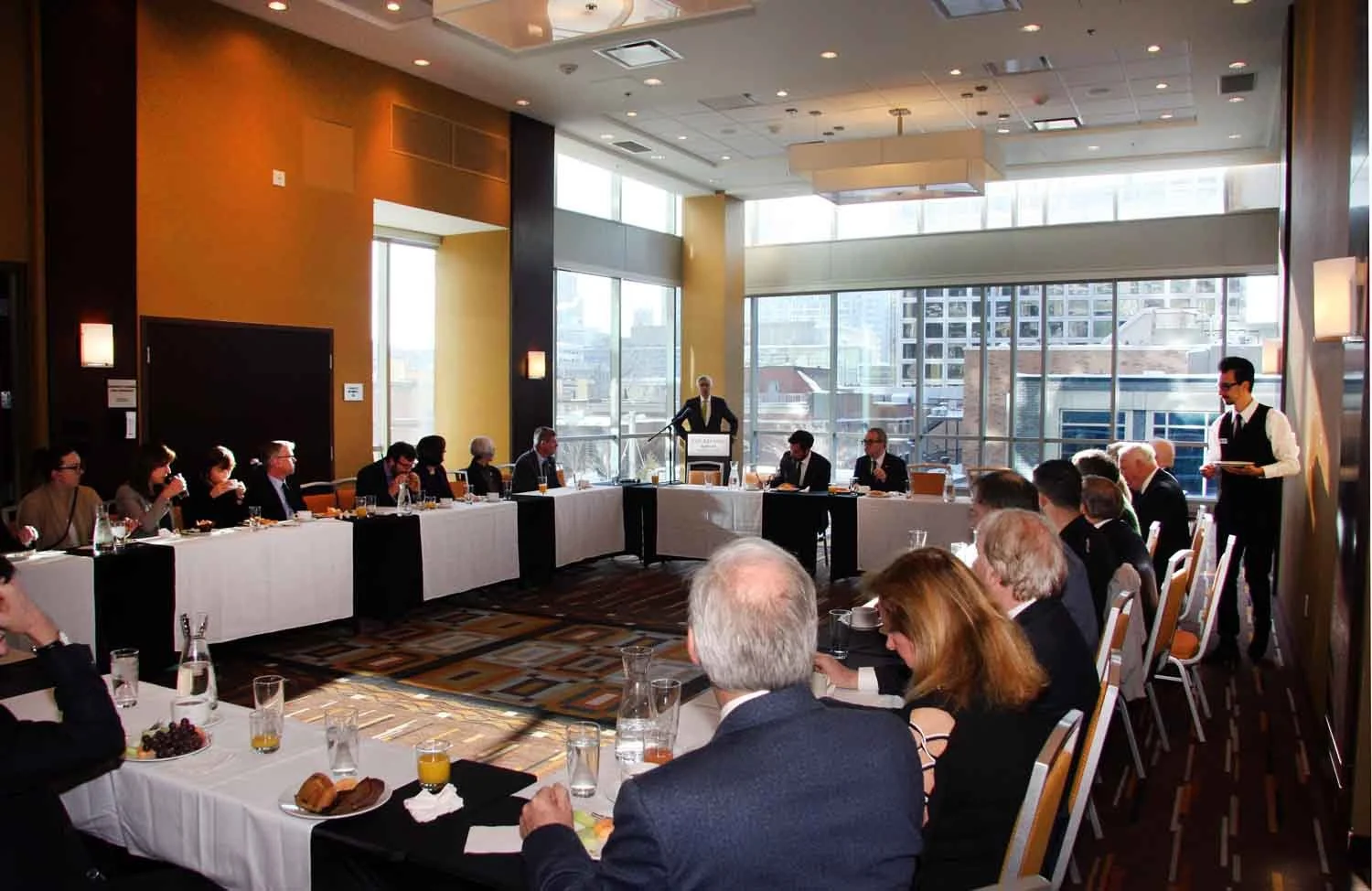 A business meeting or conference is taking place in a bright, modern room with large windows on one side. Several people are seated around tables arranged in a U-shape, listening to a speaker at a podium. Waitstaff are attending to the guests.