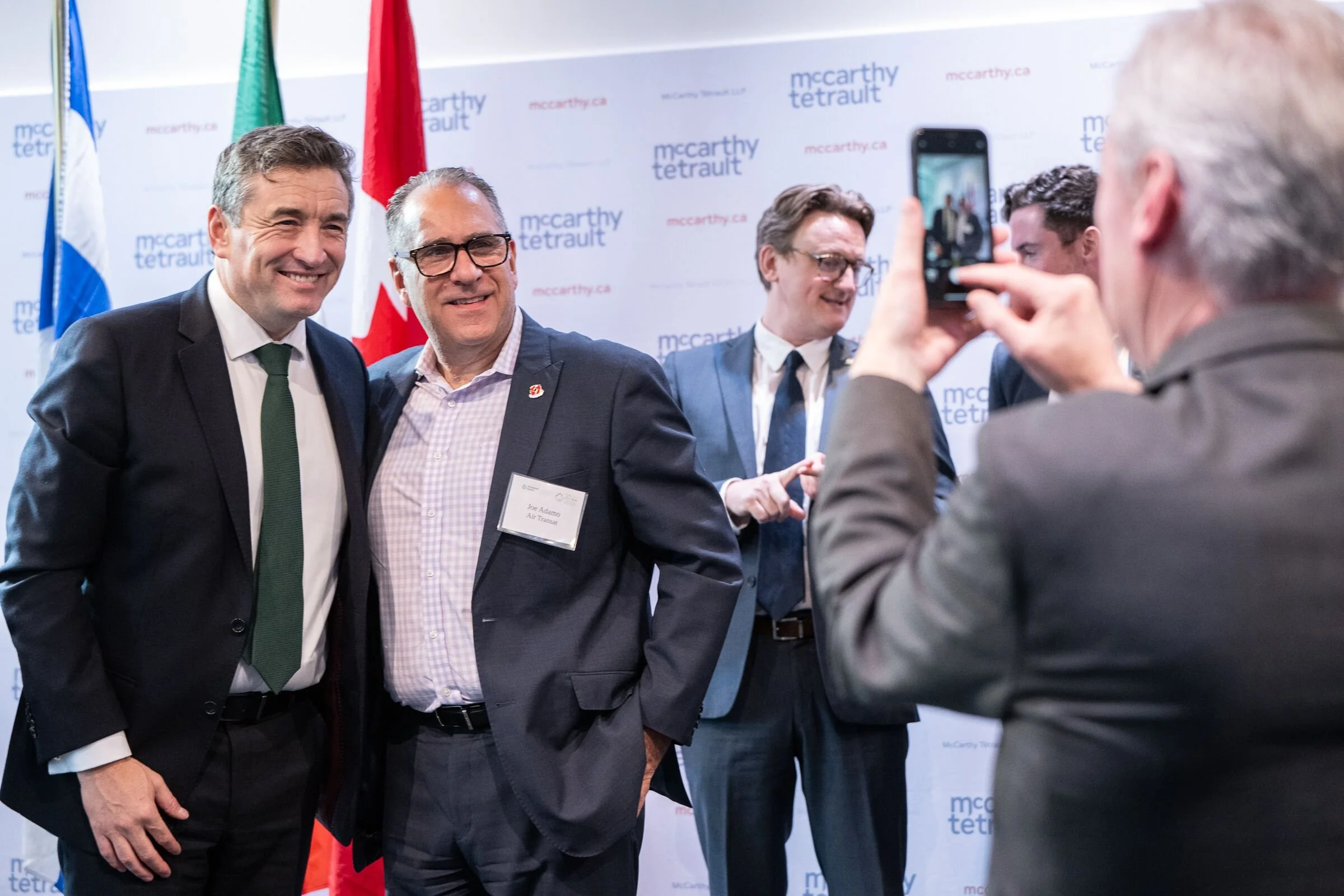 Two men in suits smiling for a photo at an event with a backdrop that reads "mccarthy tetrault." One man wears a black suit with a white shirt and green tie; the other wears a dark suit with a name tag that reads "Joe Adamo, Air Trans." Another man i