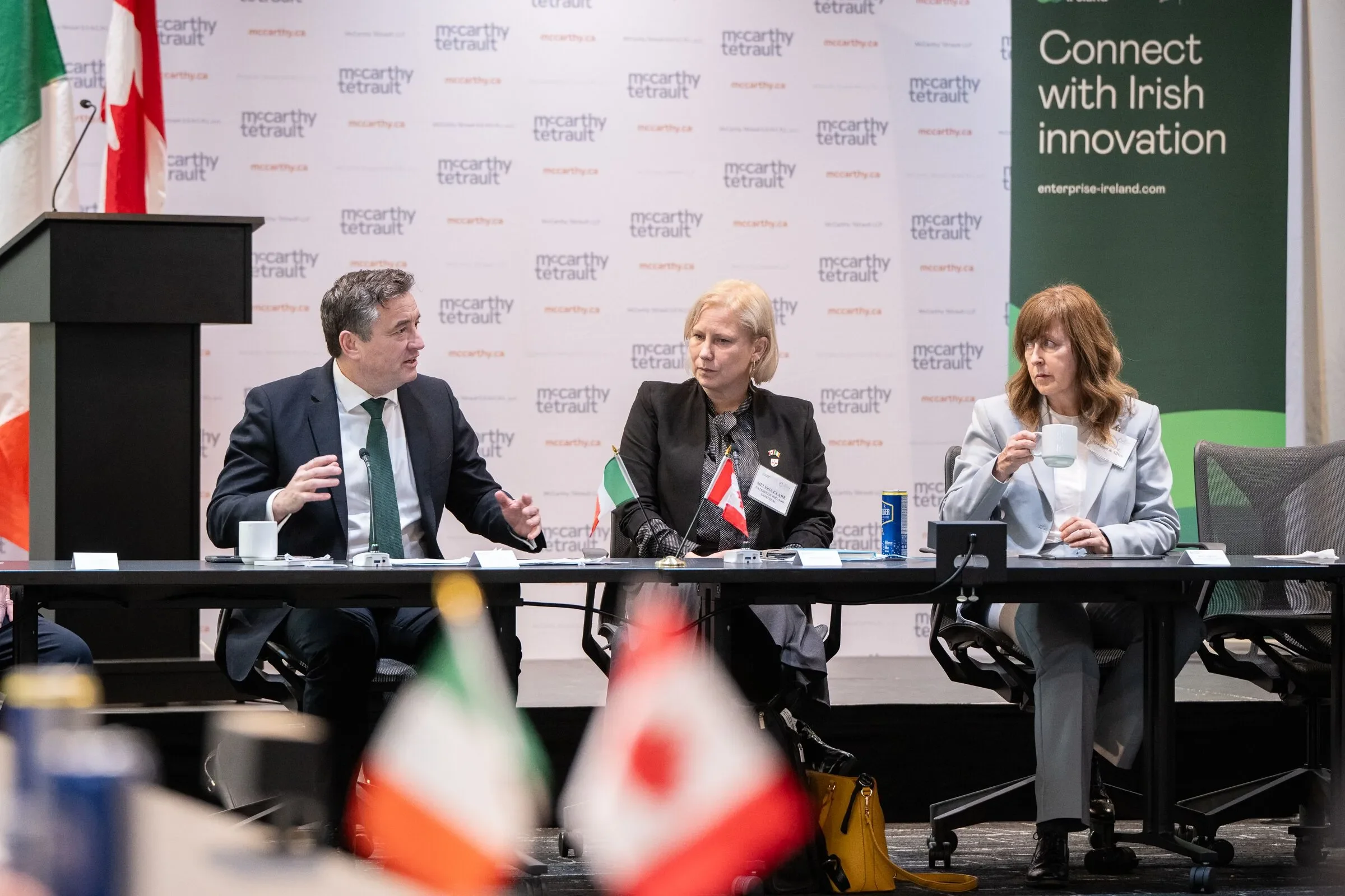 A panel of three people sitting at a conference table during a discussion at a McCarthy Tetrault event, with an Irish connection theme. One man is speaking, two women are listening, one drinking from a cup. The background has branding and banners rel