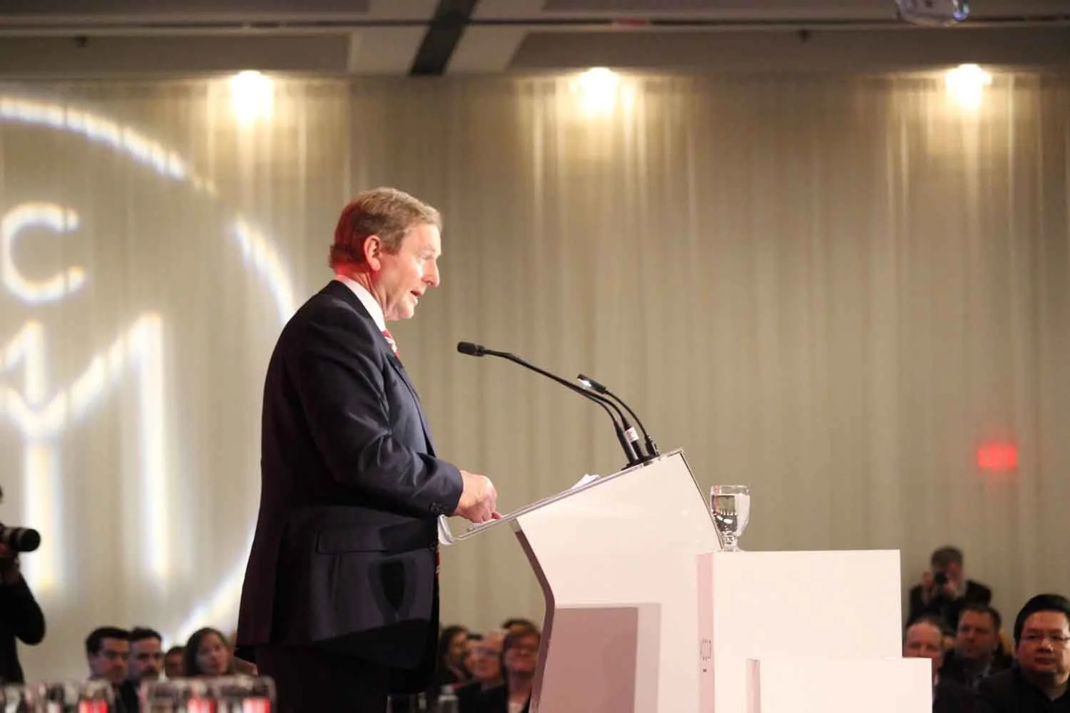A man in a dark suit giving a speech at a podium with a glass of water on it, in front of an audience in a conference hall.