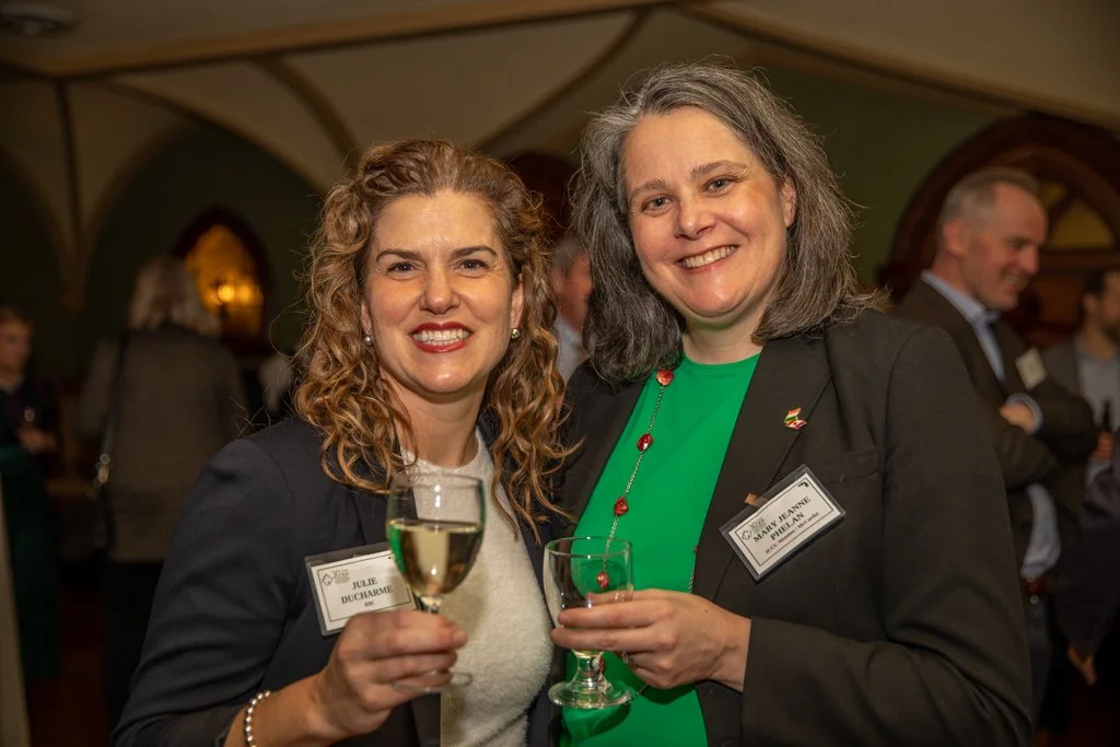 Two women smiling at a social event, holding glasses of white wine, wearing business attire with name tags, indoor setting with other people in the background.