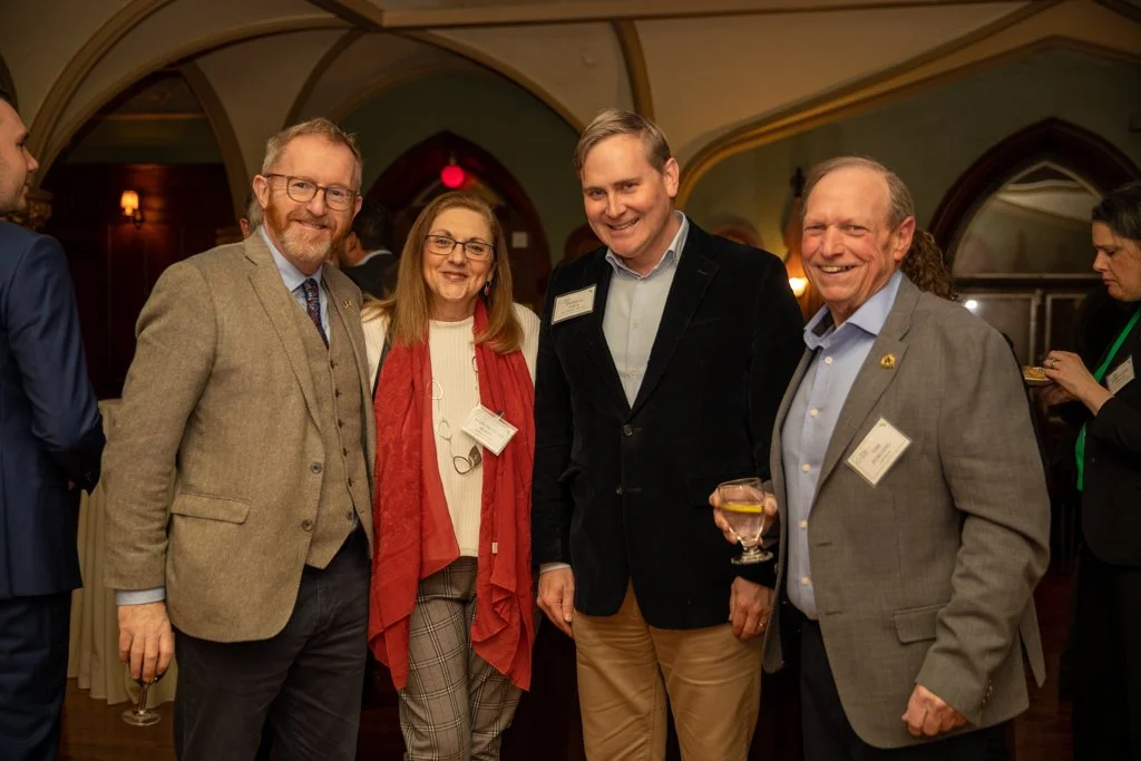 Group of five professionally dressed people at a social event, smiling and standing together in a warmly lit room with arched ceilings.