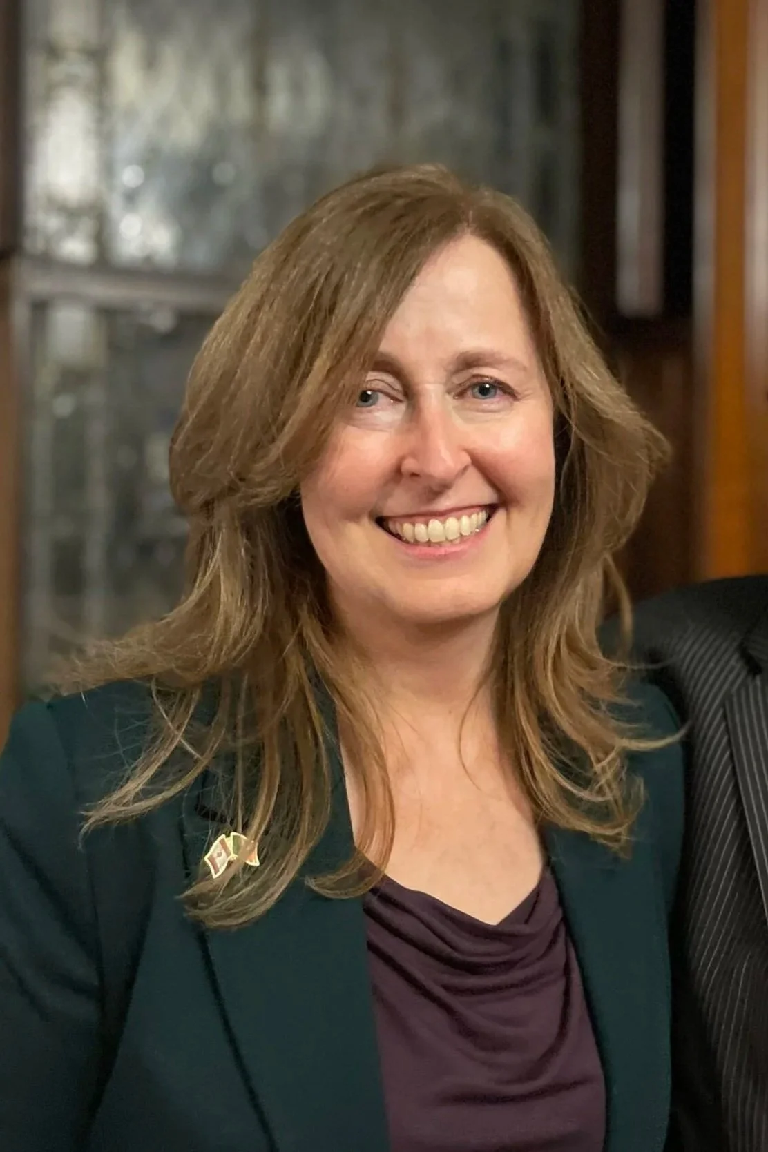 A woman with shoulder-length wavy brown hair, smiling at the camera, wearing a dark green blazer and a purple top, indoors with a wooden window frame behind her.