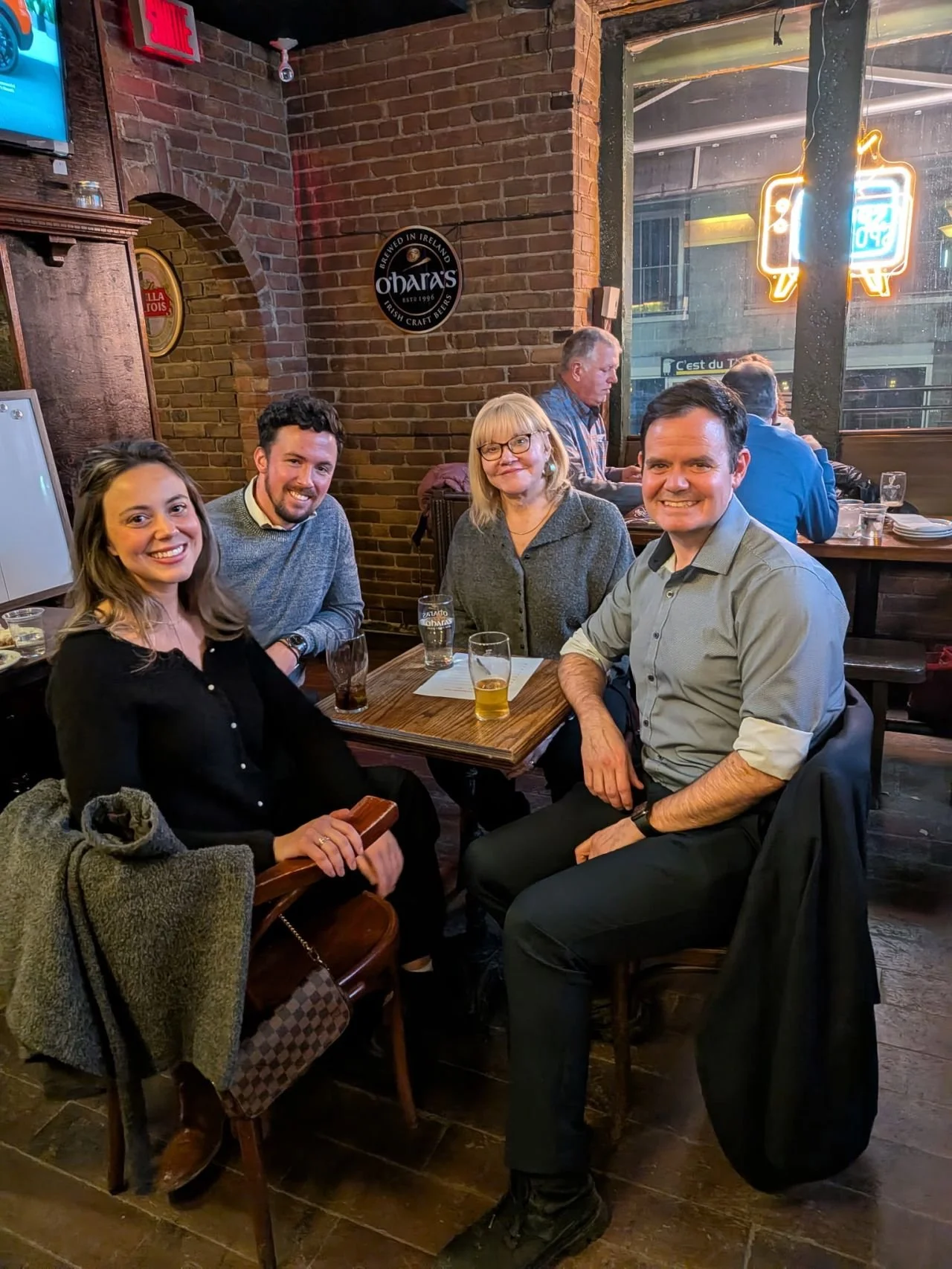 A group of four people sitting at a wooden table in a bar or restaurant, smiling at the camera. The group includes two women and two men, with drinks on the table. The background shows brick walls, a large window with neon signs, and other patrons.