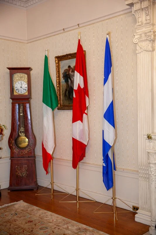 Interior of a room with a wooden grandfather clock, three flags (Italy, Canada, Greece), a framed portrait, and ornate architectural details.