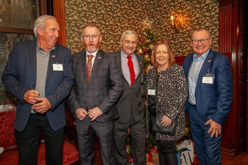 Five adults standing indoors in front of a decorated Christmas tree and wallpaper, smiling for the photo at a holiday gathering.