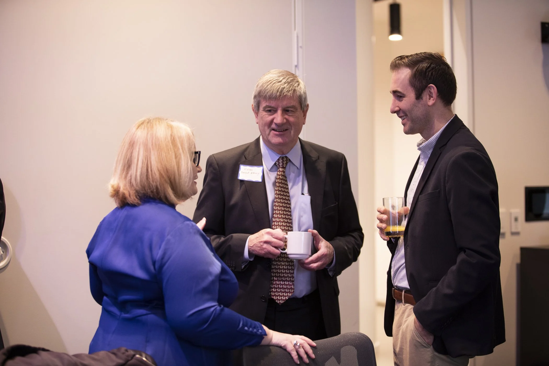 Three people engaging in conversation at a professional event, one woman and two men holding drinks, in an indoor setting.