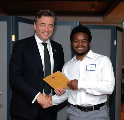 Two men shaking hands, one in a suit and the other in a white shirt, holding a yellow envelope, at an indoor event.