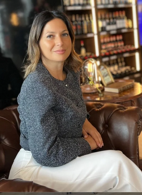 A woman sitting on a brown leather sofa in a room with wooden shelves filled with bottles in the background.