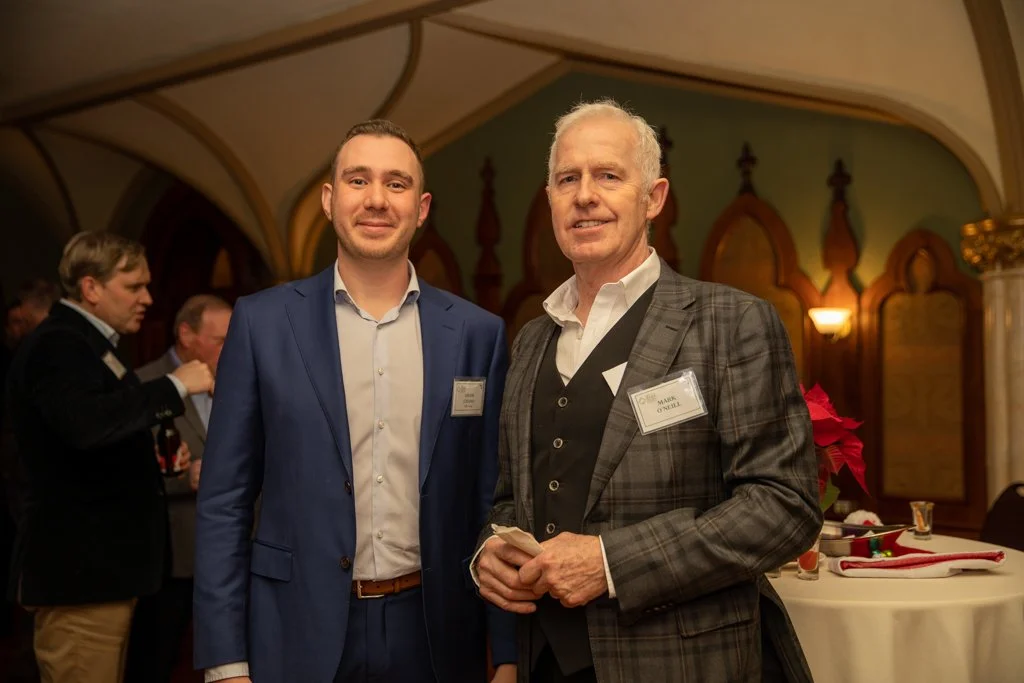 Two men in formal attire standing together at a social event, with other guests in the background, in a decorated indoor setting.