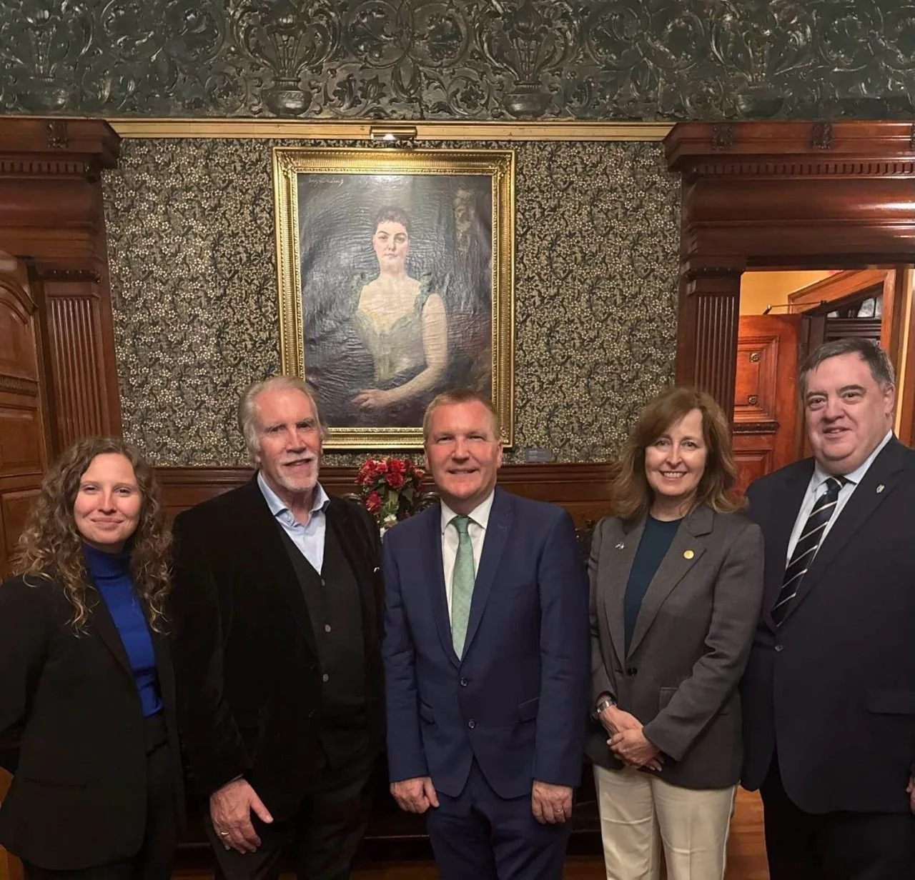 Five people standing in front of a painted portrait of a woman in a gold frame, with a dark and ornate background, inside a wood-paneled room.