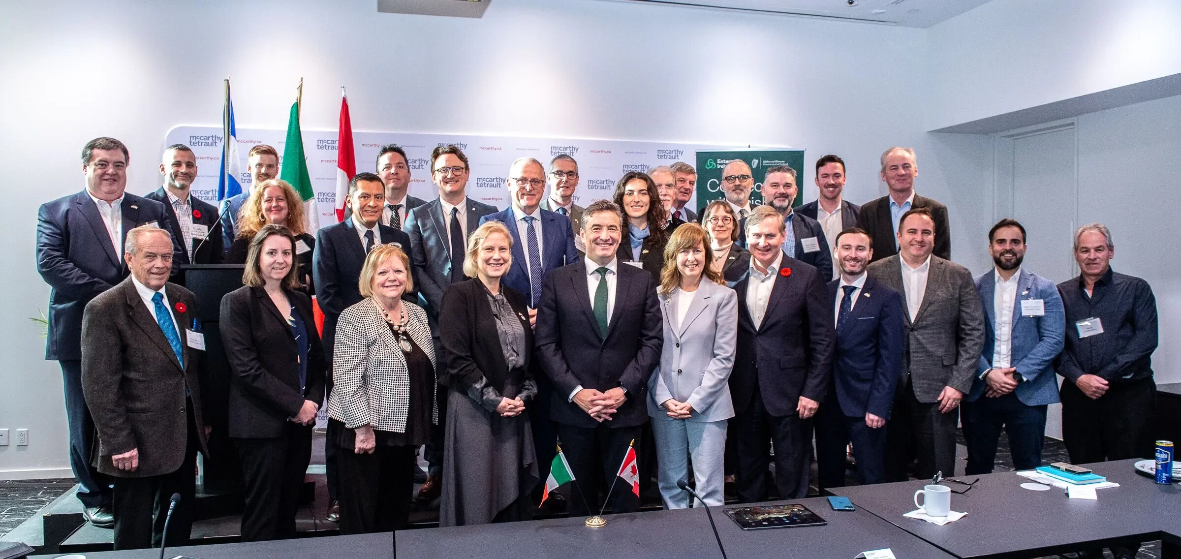 Group photo of diverse men and women in formal business attire, standing together in a conference room with flags and banners in the background.