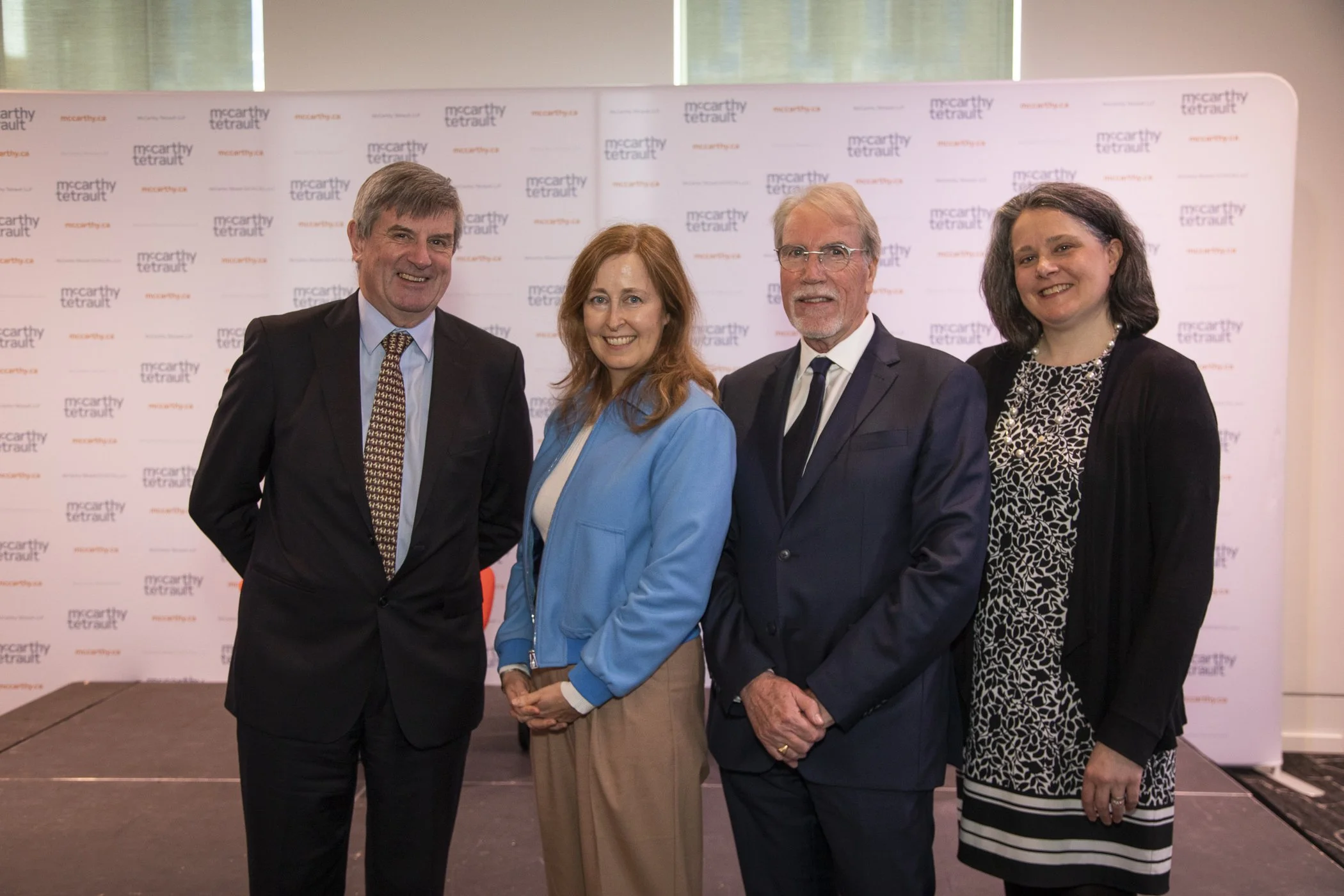 Four professionally dressed individuals standing side by side indoors, with a backdrop displaying the words "McCarthy Tetrault" repeatedly, at what appears to be a formal event or gathering.